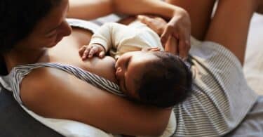 serene loving young black mother in nightgown sitting on bed and breastfeeding baby in comfortable position