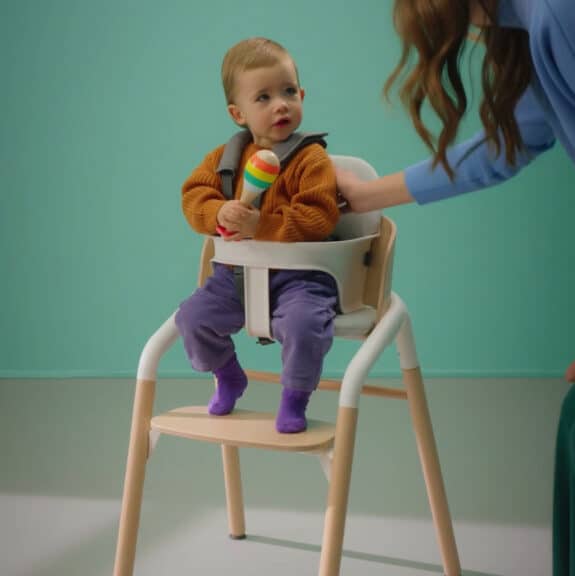 toddler in a high chair holding a toy. mom has arm on him.