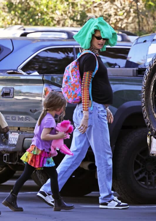 pregnant woman with backpack on and green sweater on her head holding her daughter's hand as they cross the street.
