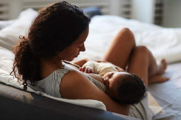 mother sitting on bed and leaning on headboard while feeding baby with breast
