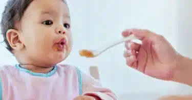 mom feeding baby girl baby food in highchair